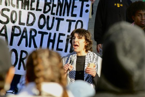 Denver School of the Arts Senior Skye, a speaker at a Nov. 9 pro-Palestine Rally at the Denver Capitol, leads chants and makes a speech for the protesters. A banner behind her accuses senator (D, CO) Michael Bennet of selling out to apartheid. High school-aged and other Gen-Z Americans like Skye have been a driving force in online advocacy for both Israel and Palestine.