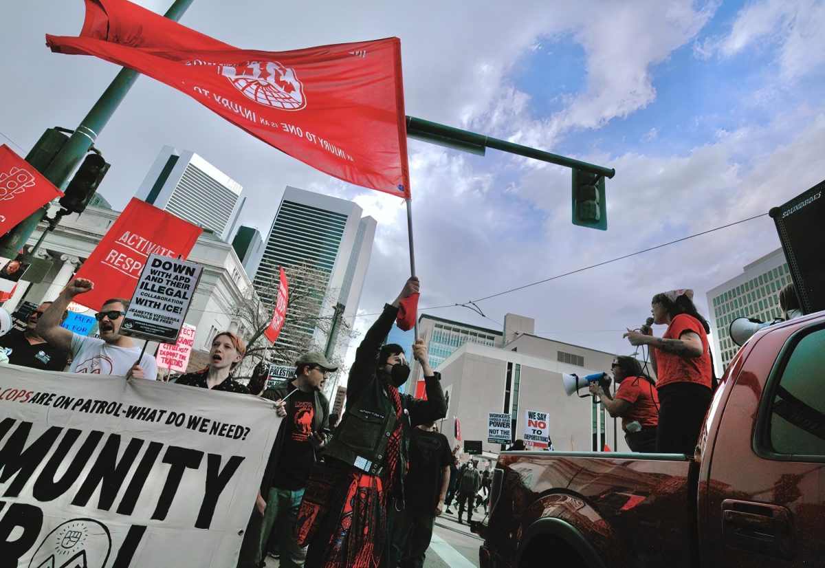 As speakers rally the crowd on the corner of 19th Street and Stout Street, a marcher hoists a flag, reading “AN INJURY TO ONE IS AN INJURY TO ALL,” a saying popularized by the Industrial Workers of the World to signify worker solidarity. The protest on May Day focused on labor unions, but also criticized the Trump administration for many grievances.